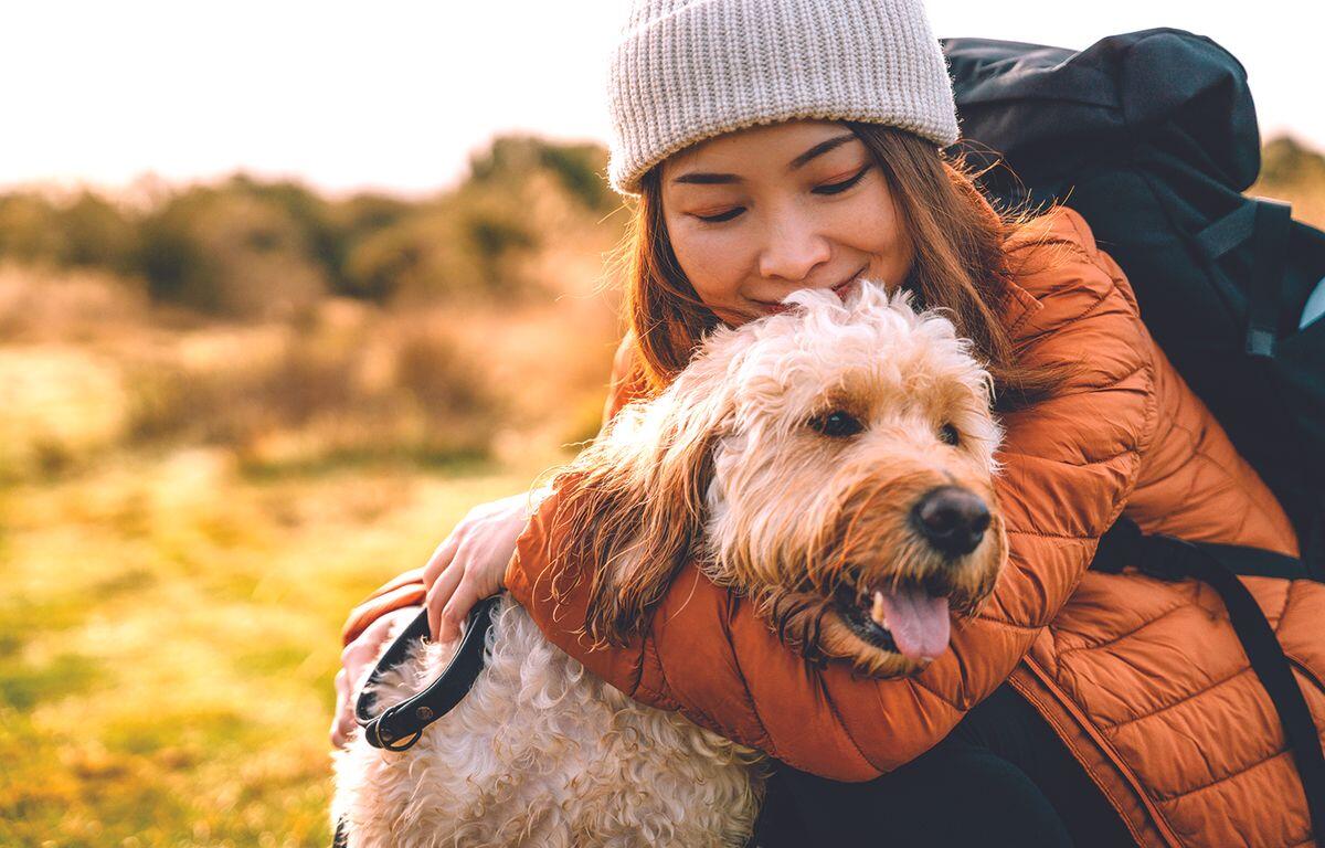 Woman in a gray beanie and orange puffer jacket hugs a fluffy light-brown dog outdoors in a sunlit field.