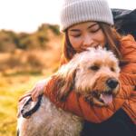 Woman in a gray beanie and orange puffer jacket hugs a fluffy light-brown dog outdoors in a sunlit field.