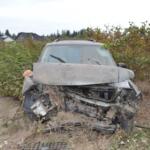 Front end of a crushed SUV with a bent hood and exposed engine, sitting in a dirt lot surrounded by bushes and vines in a rural area