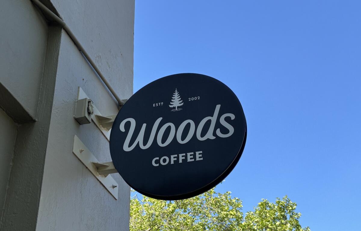 Dark blue circular Woods Coffee sign with white script and a small tree logo, mounted on a light building under a clear blue sky
