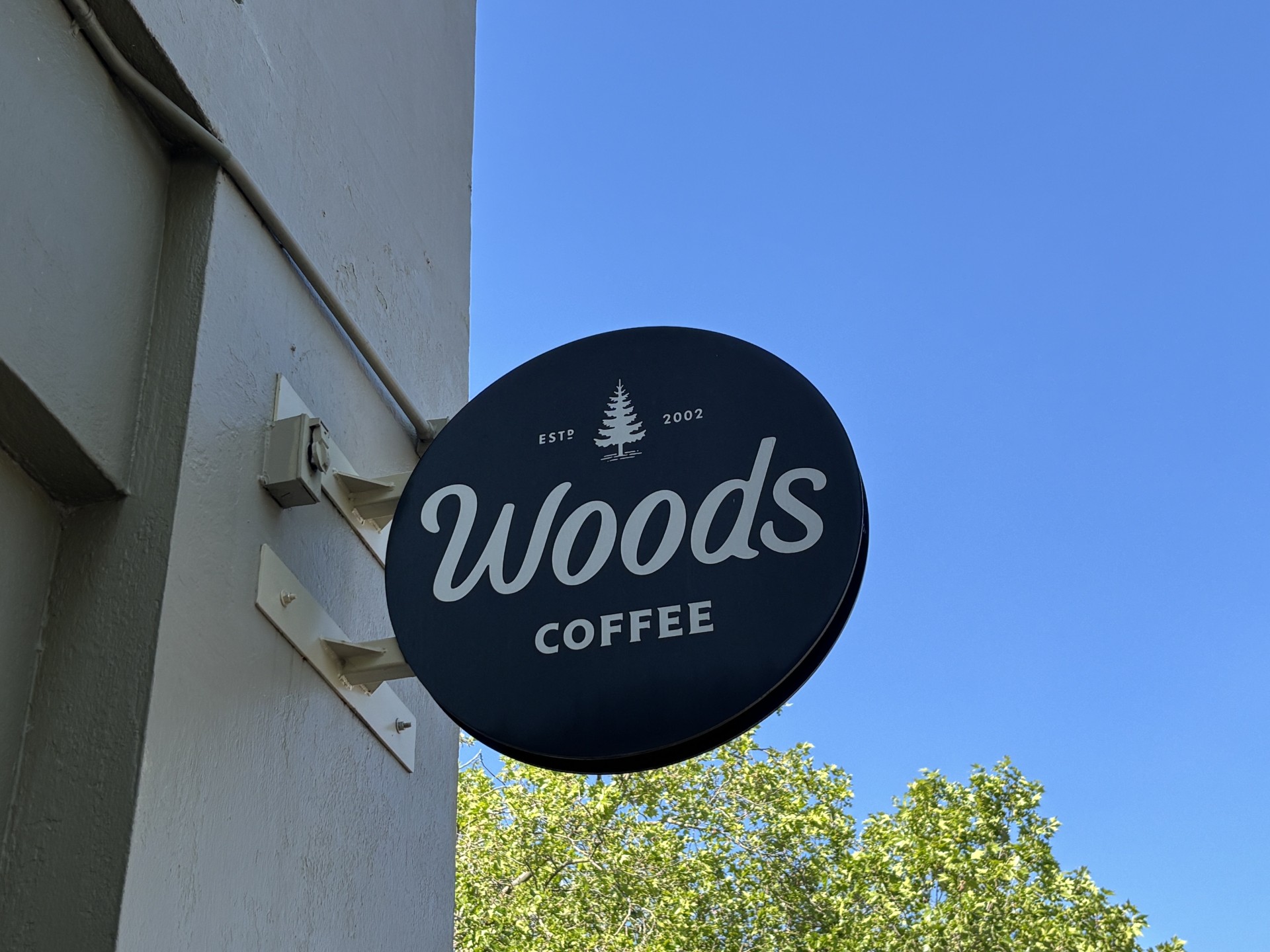 Dark blue circular Woods Coffee sign with white script and a small tree logo, mounted on a light building under a clear blue sky