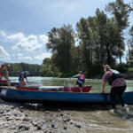 People launch onto a calm river from a rocky shore, wearing life jackets and helmets beside several canoes under a blue sky with trees in the background.