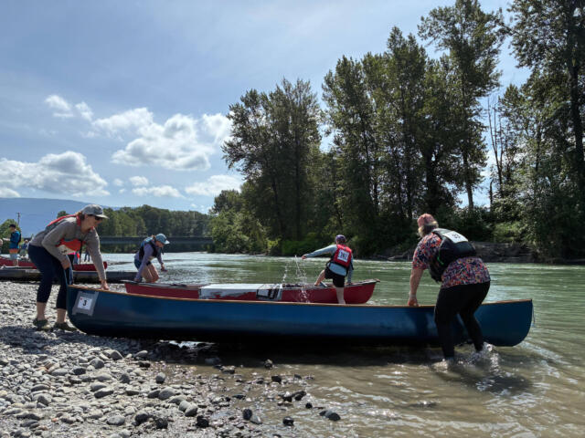 People launch onto a calm river from a rocky shore, wearing life jackets and helmets beside several canoes under a blue sky with trees in the background.
