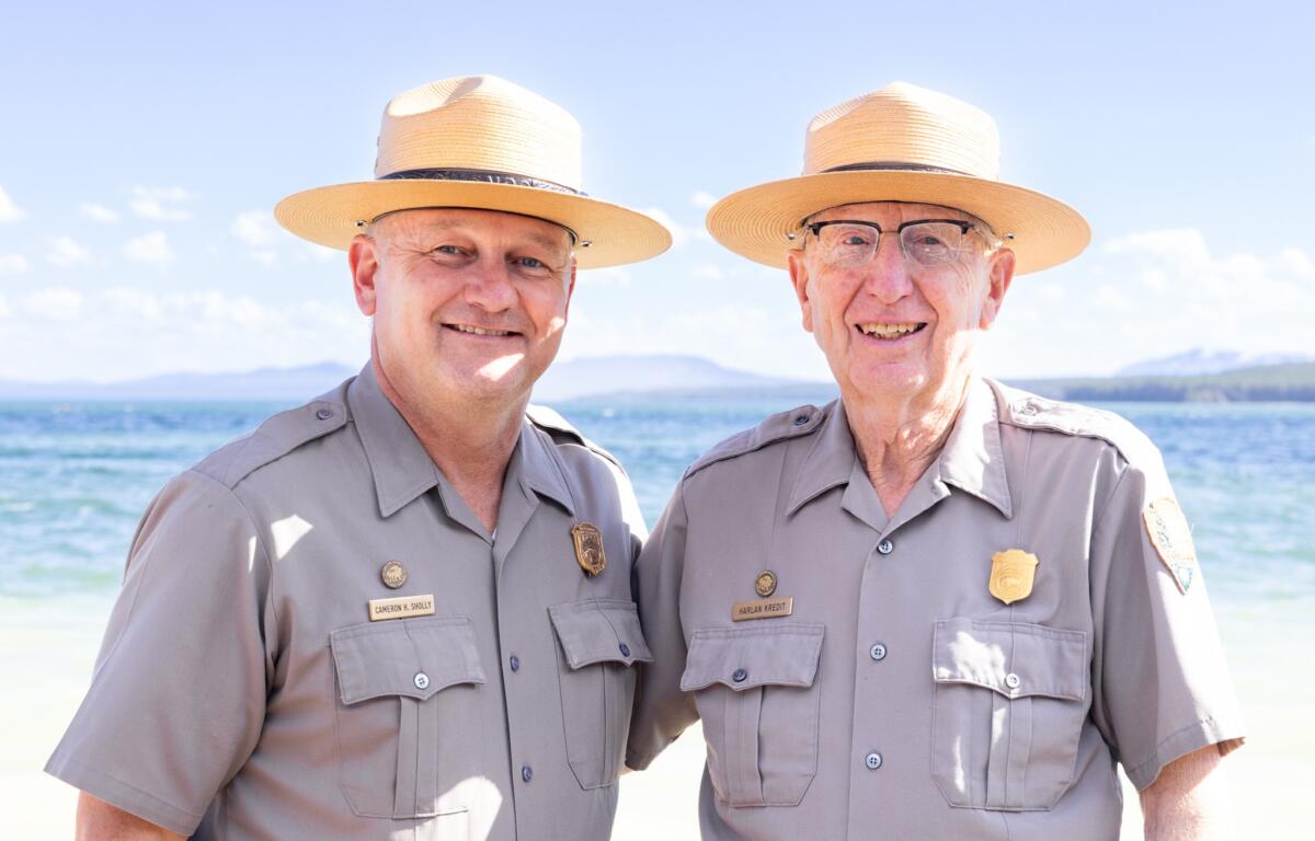 Two men in khaki uniforms and wide-brimmed straw hats stand close together on a sunny beach, smiling at the camera with water in the background.