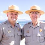 Two men in khaki uniforms and wide-brimmed straw hats stand close together on a sunny beach, smiling at the camera with water in the background.