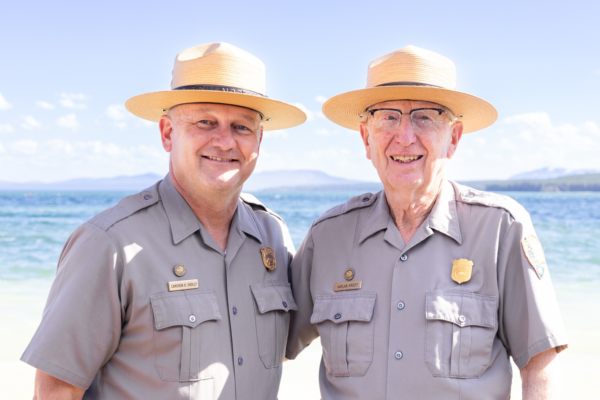 Two men in khaki uniforms and wide-brimmed straw hats stand close together on a sunny beach, smiling at the camera with water in the background.