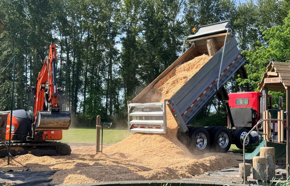Orange excavator dumping a load of sand into a tilted dump trailer at a construction site, with trees in the background.
