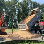 Orange excavator dumping a load of sand into a tilted dump trailer at a construction site, with trees in the background.