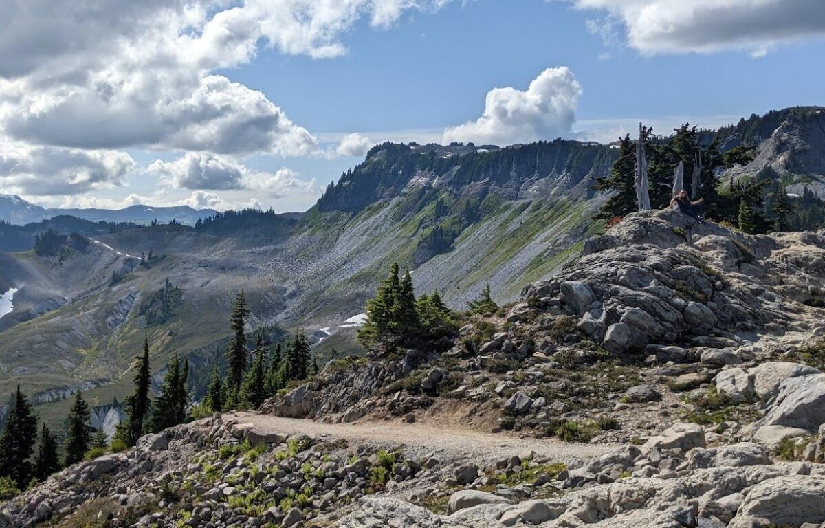 Two hikers sit on a rocky outcrop on a mountain cliff, with a valley and pine trees below under a cloudy sky.