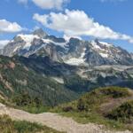 Snowy, jagged mountain peak under a blue sky, viewed from a rocky dirt trail with green shrubs in the foreground.