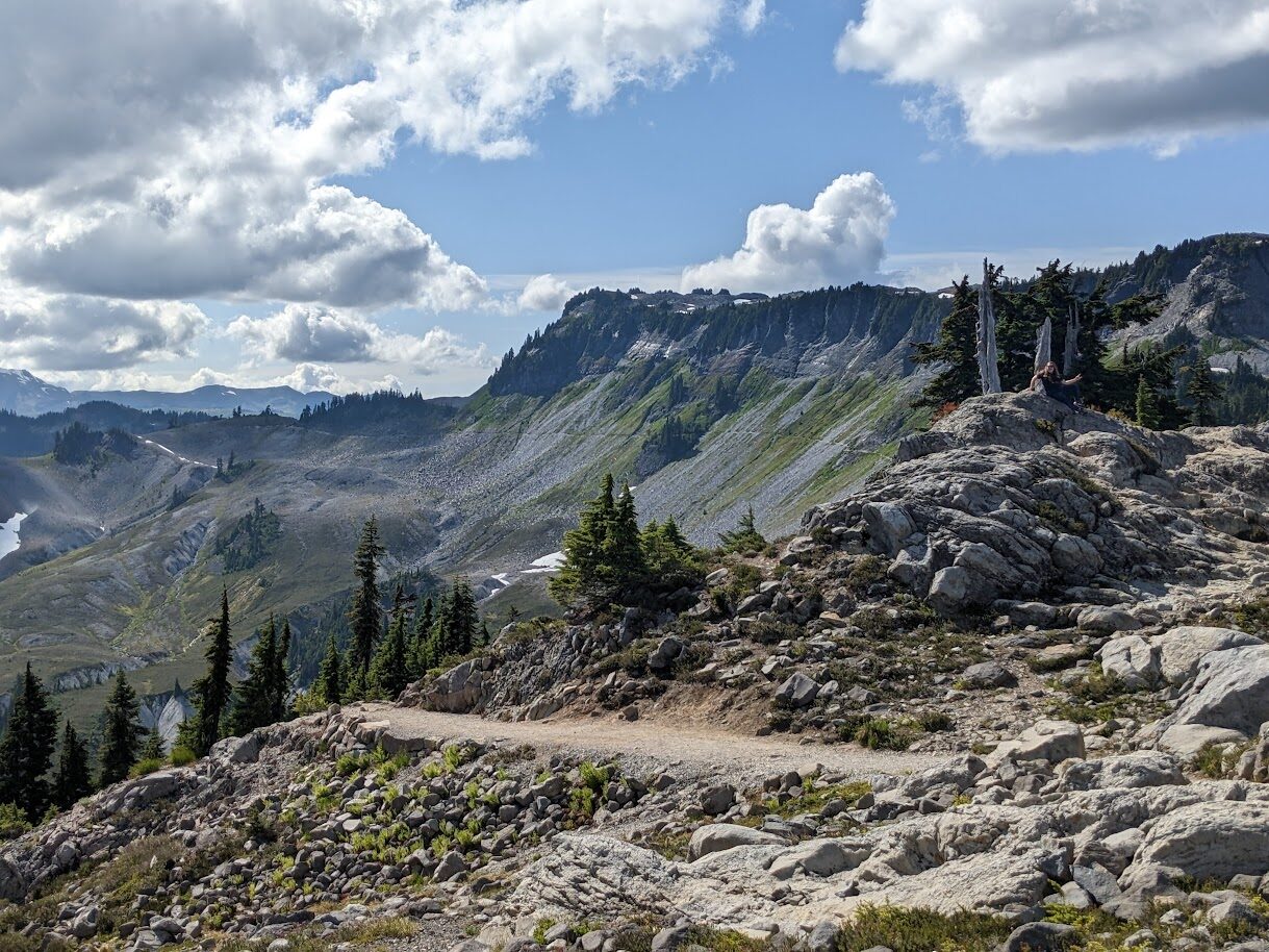 Two hikers sit on a rocky outcrop on a mountain cliff, with a valley and pine trees below under a cloudy sky.