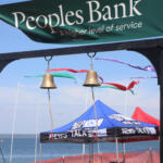 Green banner reading 'Peoples Bank' above two hanging bells at a lakeside event, with colorful tents and blue sky in the background.