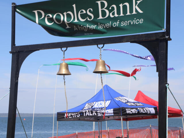 Green banner reading 'Peoples Bank' above two hanging bells at a lakeside event, with colorful tents and blue sky in the background.