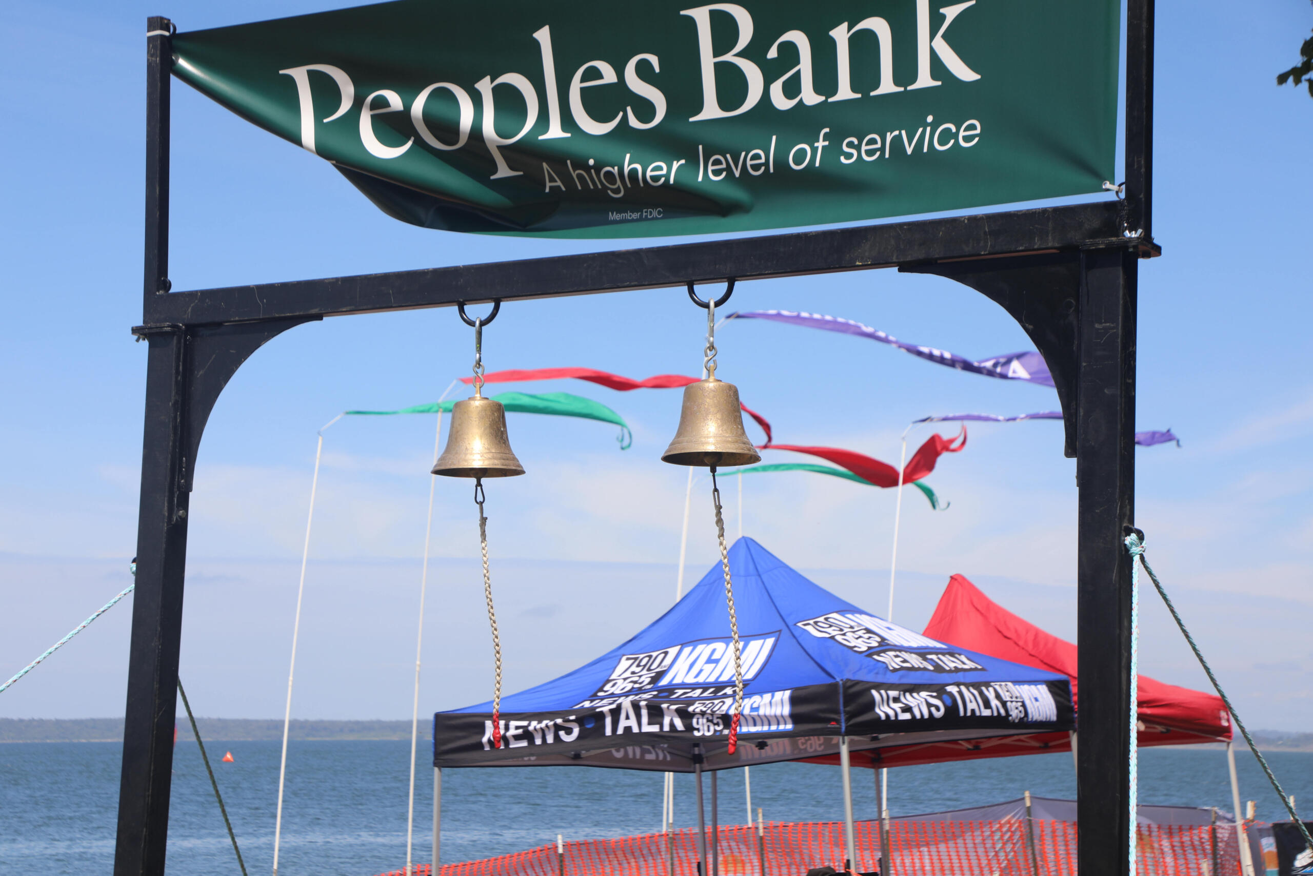 Green banner reading 'Peoples Bank' above two hanging bells at a lakeside event, with colorful tents and blue sky in the background.