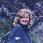 Smiling woman with blond hair wearing a dark jacket and backpack outdoors beside a rocky cliff.