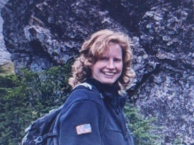 Smiling woman with blond hair wearing a dark jacket and backpack outdoors beside a rocky cliff.