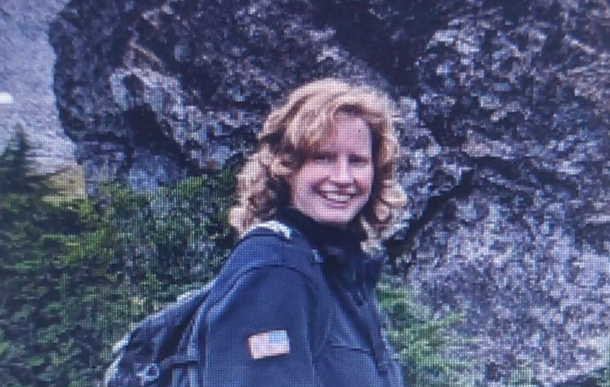 Smiling woman with blond hair wearing a dark jacket and backpack outdoors beside a rocky cliff.