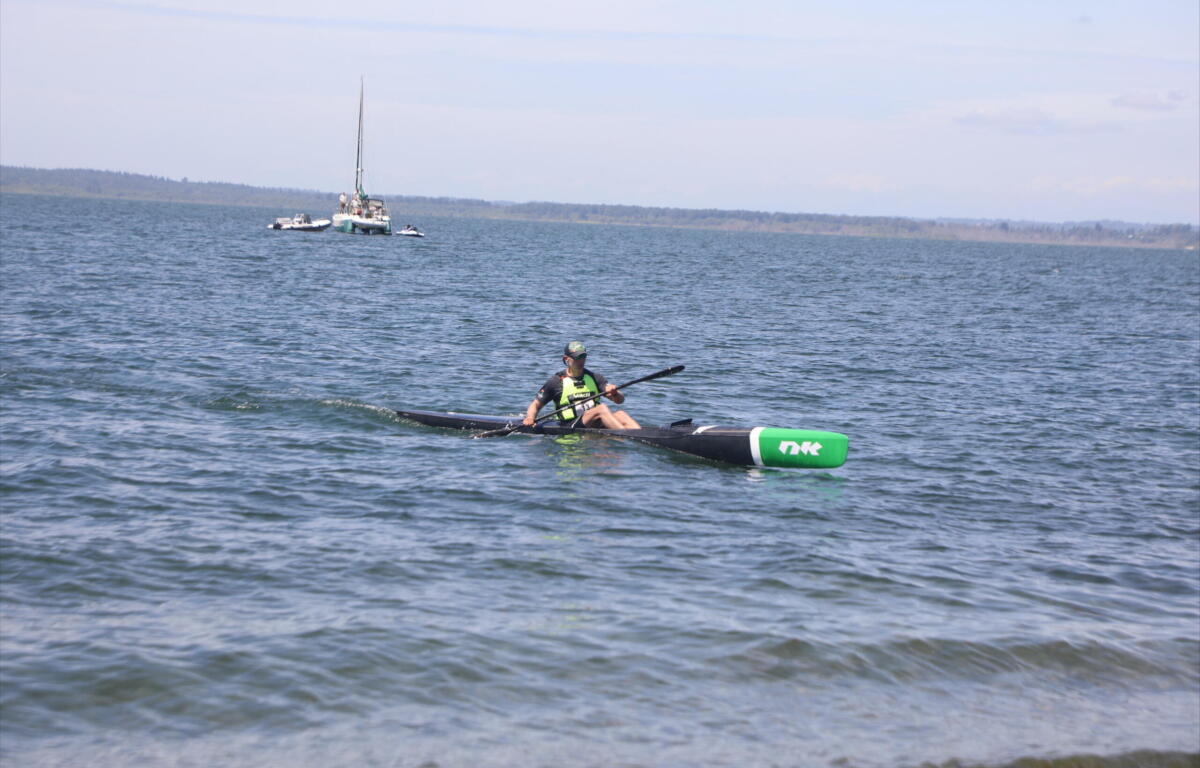 A kayaker in a green life jacket paddles a long black kayak on a calm lake with boats anchored in the distance and a shoreline on the horizon.