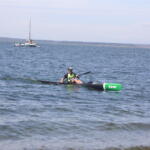 A kayaker in a green life jacket paddles a long black kayak on a calm lake with boats anchored in the distance and a shoreline on the horizon.