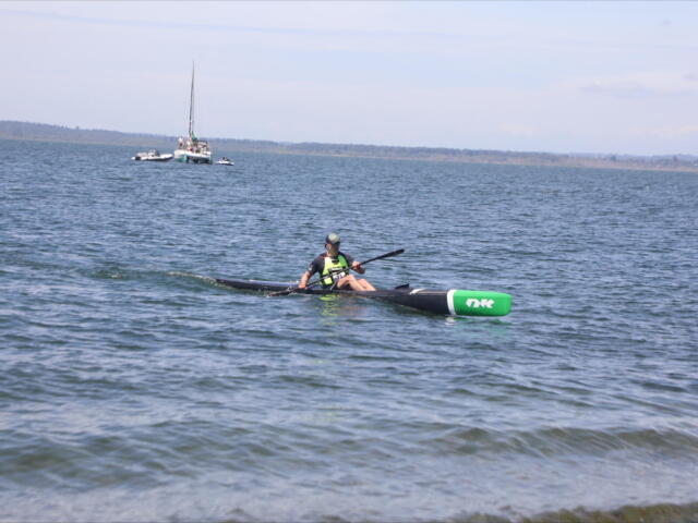 A kayaker in a green life jacket paddles a long black kayak on a calm lake with boats anchored in the distance and a shoreline on the horizon.