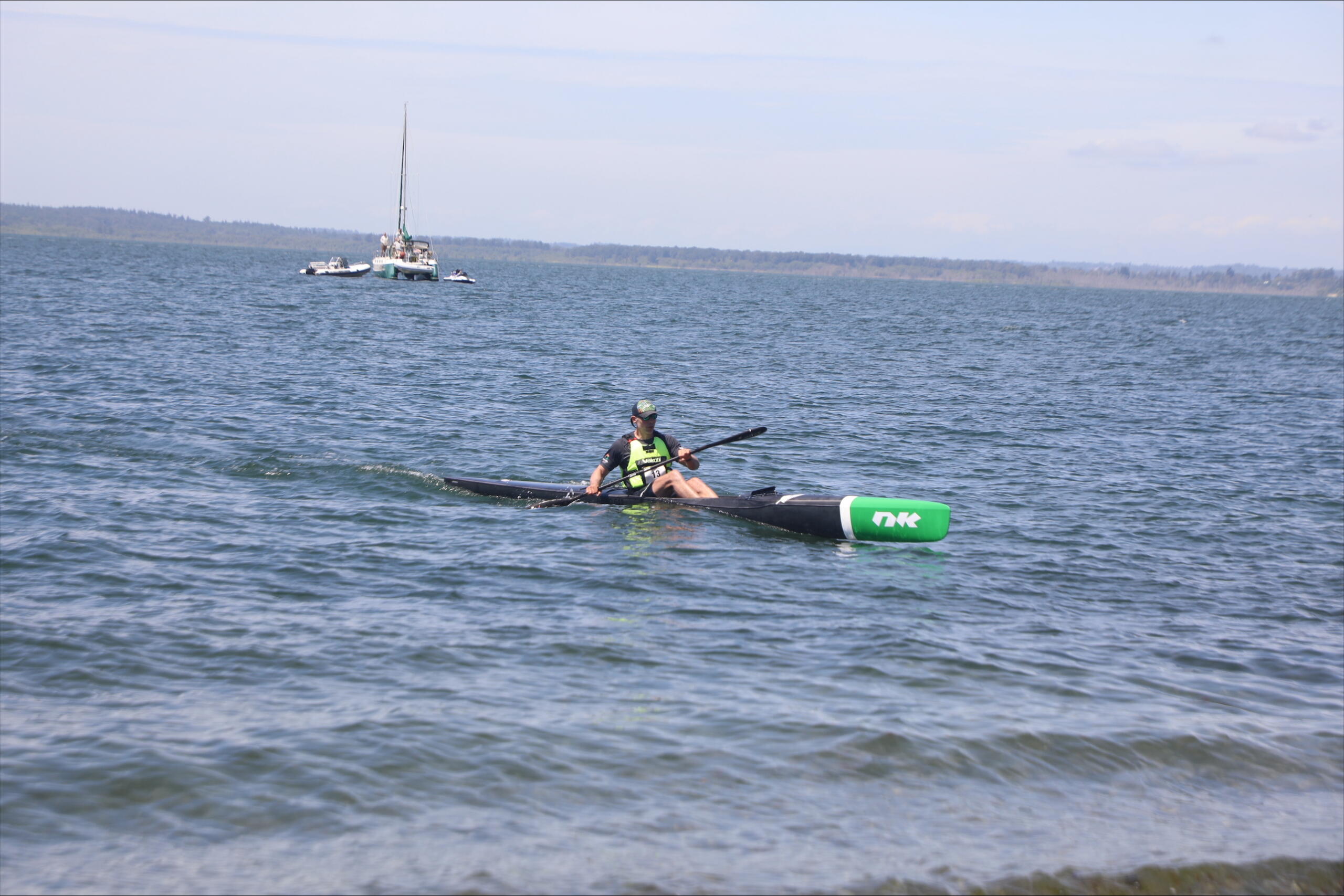 A kayaker in a green life jacket paddles a long black kayak on a calm lake with boats anchored in the distance and a shoreline on the horizon.