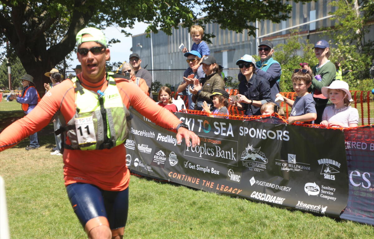 Runner in orange athletic gear crosses a finish line as a cheering crowd and sponsor banner loom in the background.