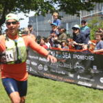 Runner in orange athletic gear crosses a finish line as a cheering crowd and sponsor banner loom in the background.