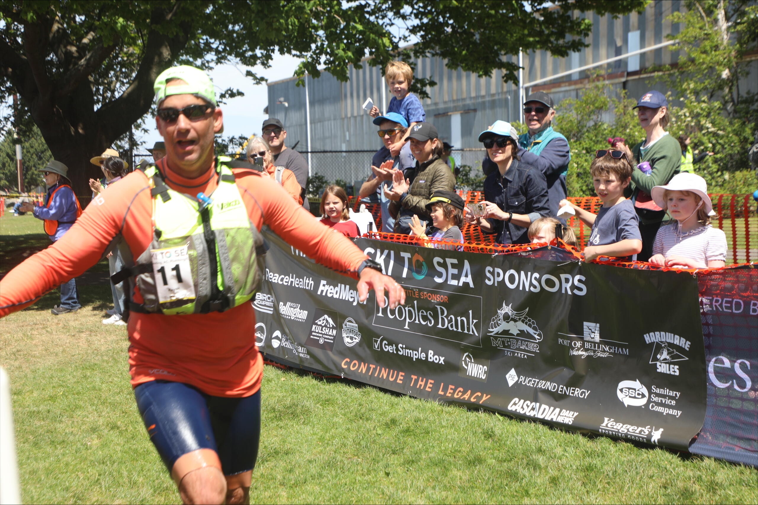 Runner in orange athletic gear crosses a finish line as a cheering crowd and sponsor banner loom in the background.