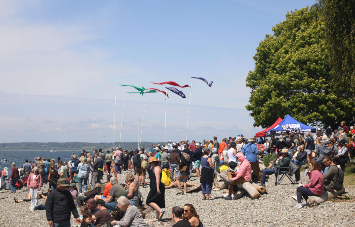Large crowd gathered on a pebbled beach for an outdoor event, with colorful flags on tall poles and tents to the right.