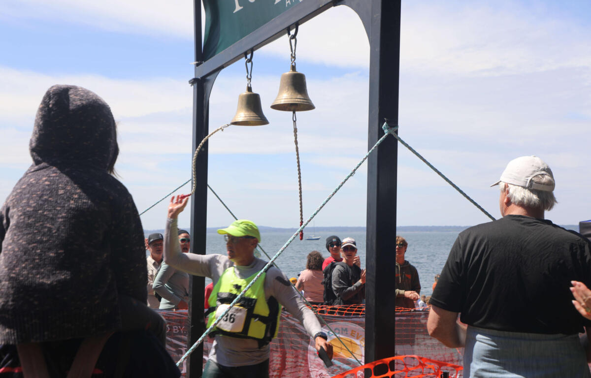 Crowd of runners and spectators at a seaside race finish under hanging bells on a metal frame.