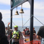 Crowd of runners and spectators at a seaside race finish under hanging bells on a metal frame.