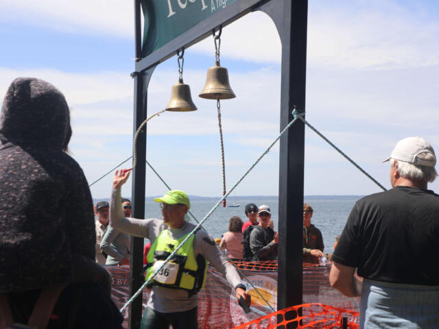 Crowd of runners and spectators at a seaside race finish under hanging bells on a metal frame.