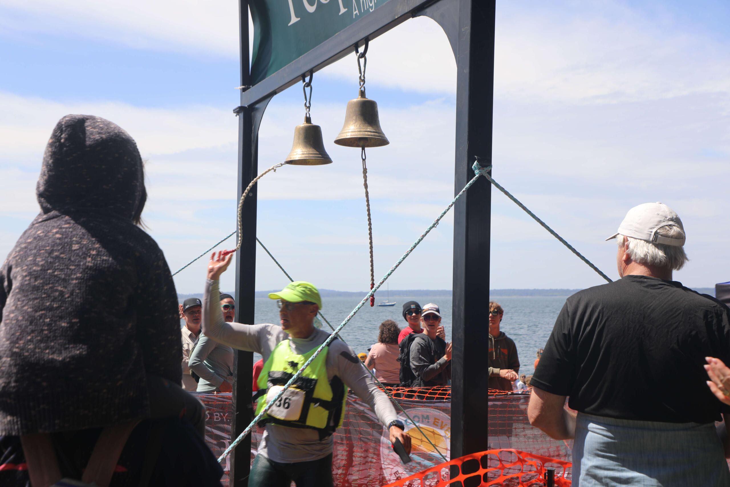 Crowd of runners and spectators at a seaside race finish under hanging bells on a metal frame.