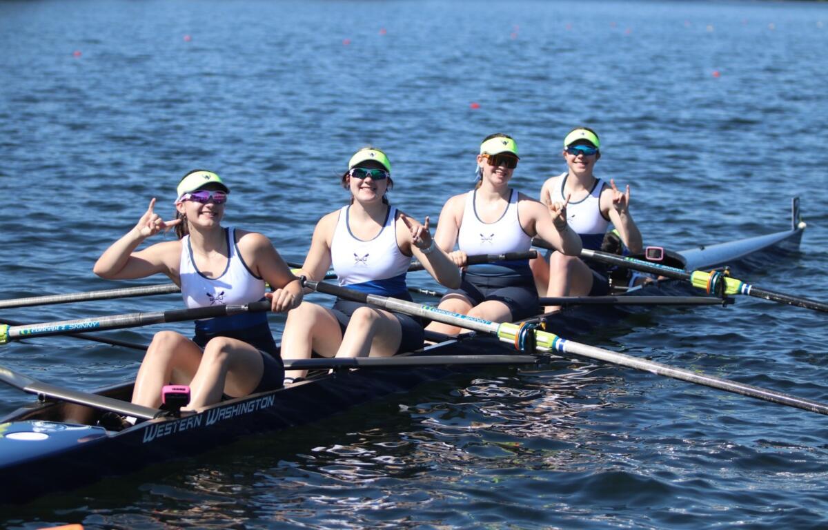 Four female rowers in a white-and-navy shell on a blue lake, posing with rock-on hand signs after a stroke, wearing neon visors and sunglasses.
