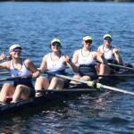 Four female rowers in a white-and-navy shell on a blue lake, posing with rock-on hand signs after a stroke, wearing neon visors and sunglasses.