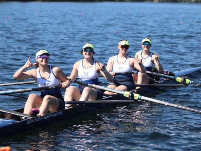 Four female rowers in a white-and-navy shell on a blue lake, posing with rock-on hand signs after a stroke, wearing neon visors and sunglasses.