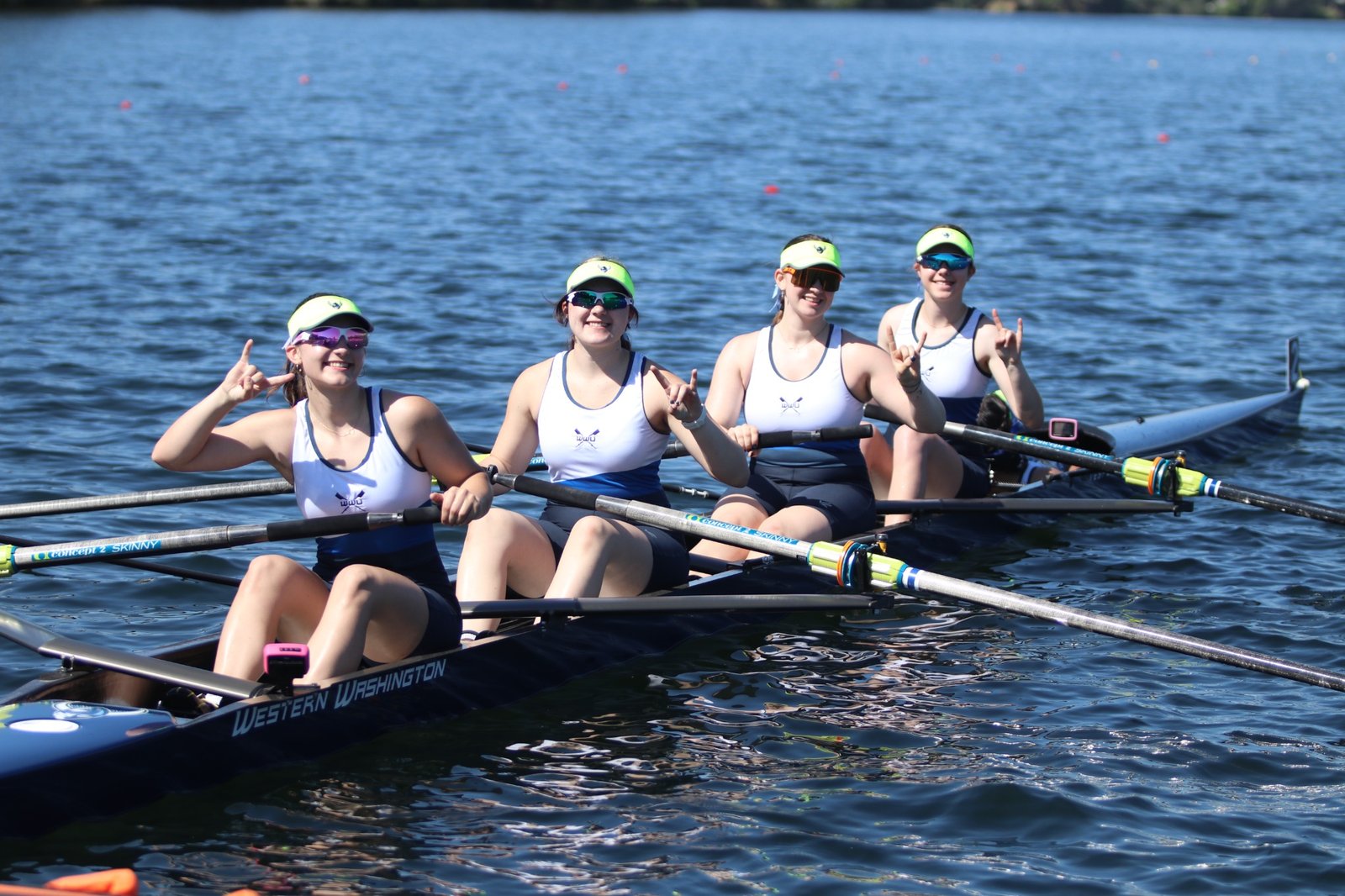 Four female rowers in a white-and-navy shell on a blue lake, posing with rock-on hand signs after a stroke, wearing neon visors and sunglasses.