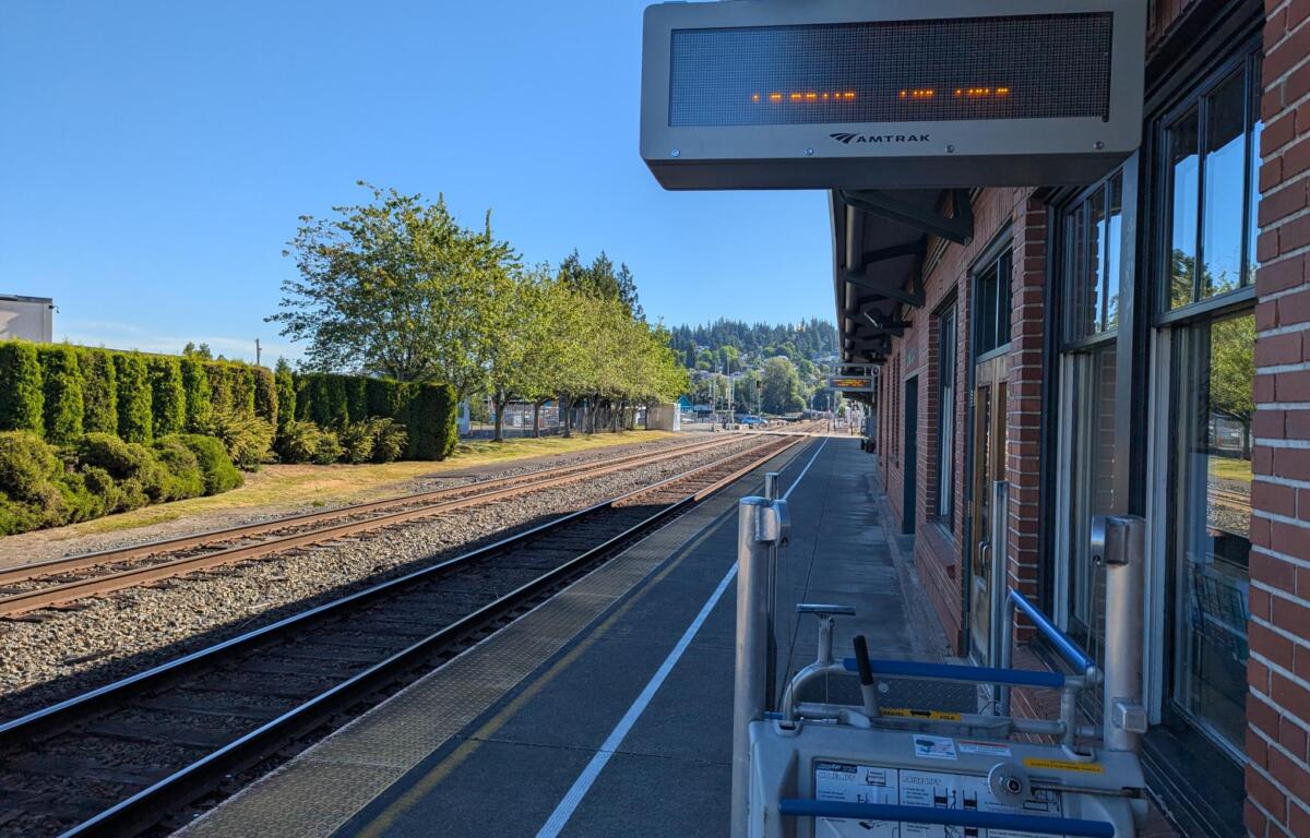 Amtrak platform with brick building, safety railing, and overhead electronic sign above tracks on a sunny day.