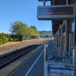 Amtrak platform with brick building, safety railing, and overhead electronic sign above tracks on a sunny day.