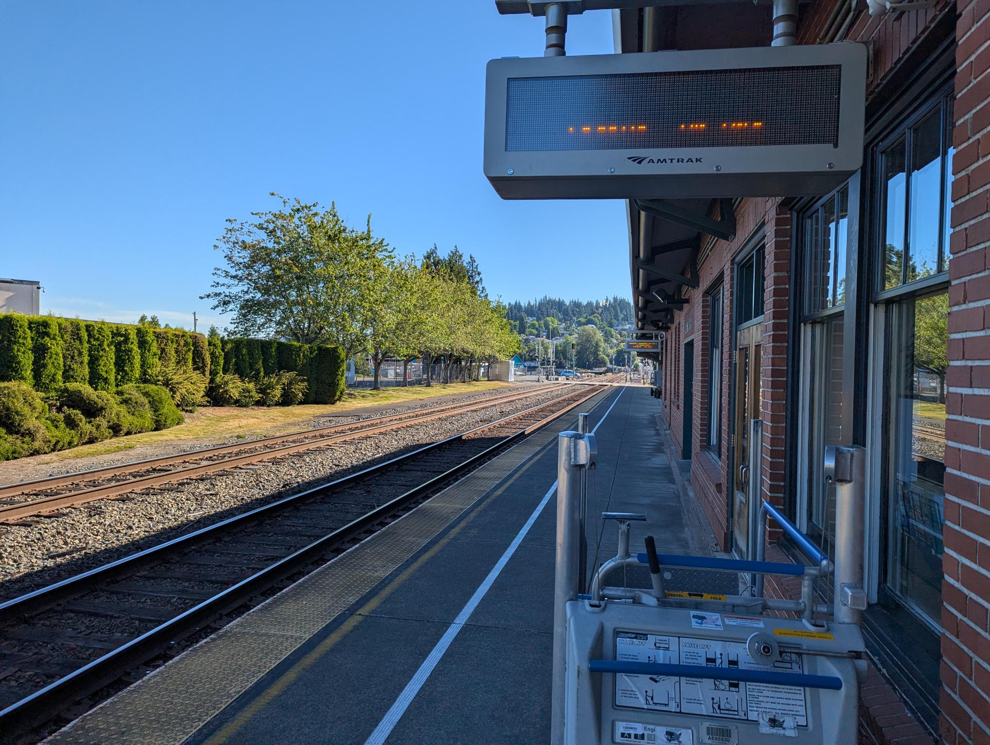Amtrak platform with brick building, safety railing, and overhead electronic sign above tracks on a sunny day.