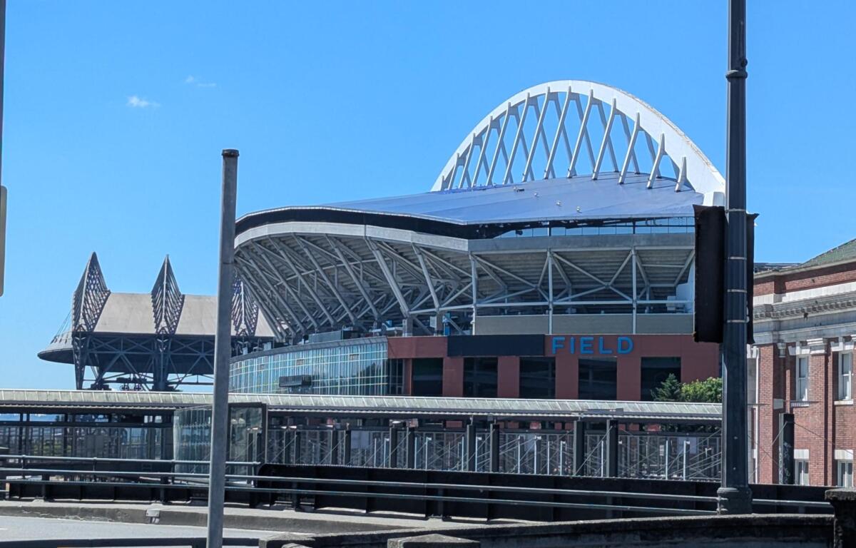Exterior view of a stadium with a white arched lattice roof against a blue sky, visible seating and glass facade.