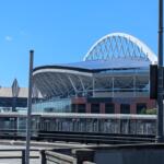 Exterior view of a stadium with a white arched lattice roof against a blue sky, visible seating and glass facade.