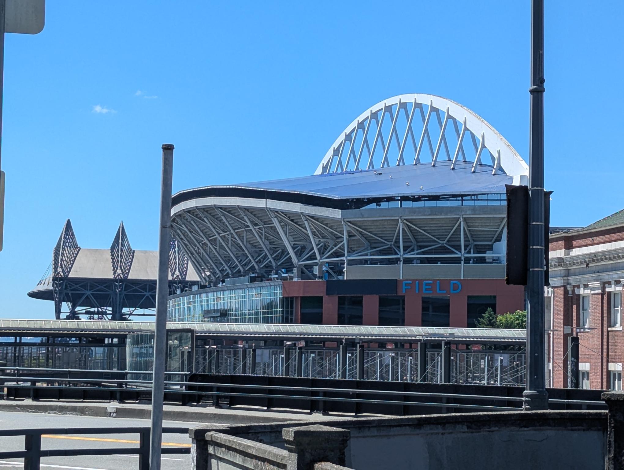 Exterior view of a stadium with a white arched lattice roof against a blue sky, visible seating and glass facade.