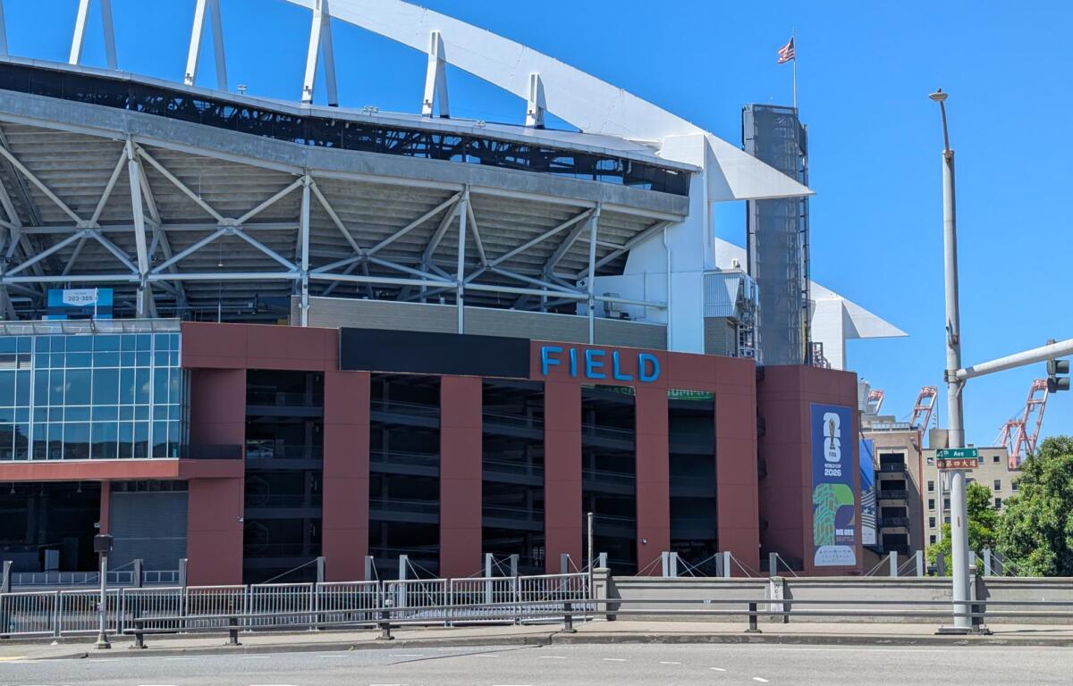 Exterior of a stadium with white steel framework and a red-brown façade, blue 'FIELD' lettering visible under a clear blue sky