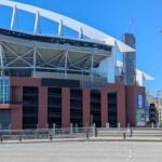 Exterior of a stadium with white steel framework and a red-brown façade, blue 'FIELD' lettering visible under a clear blue sky