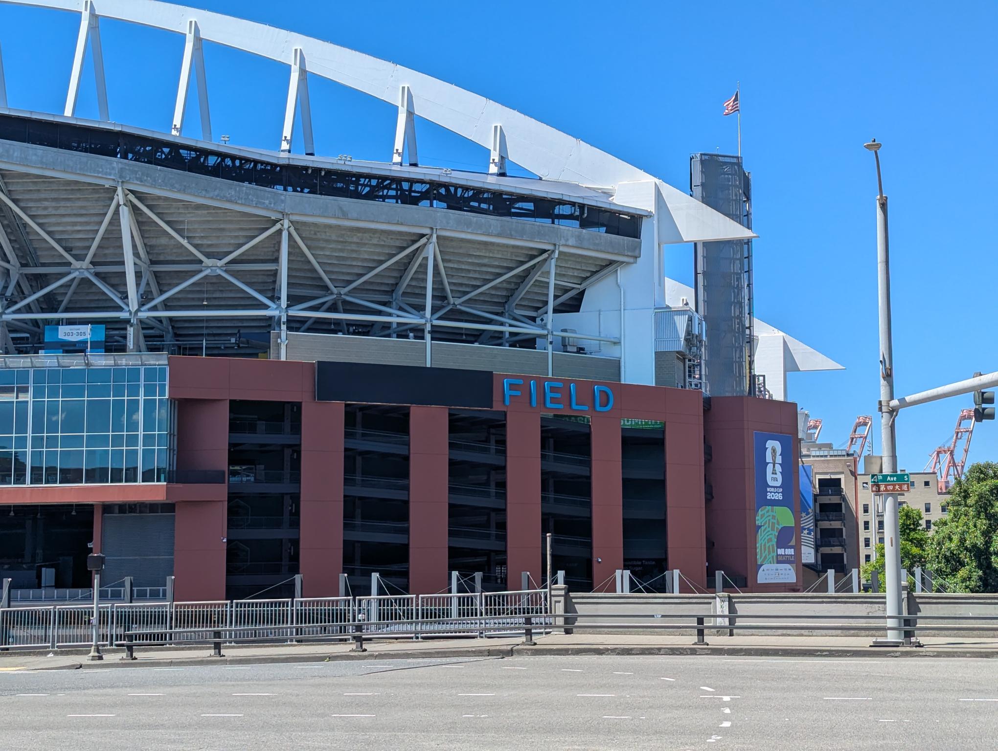 Exterior of a stadium with white steel framework and a red-brown façade, blue 'FIELD' lettering visible under a clear blue sky