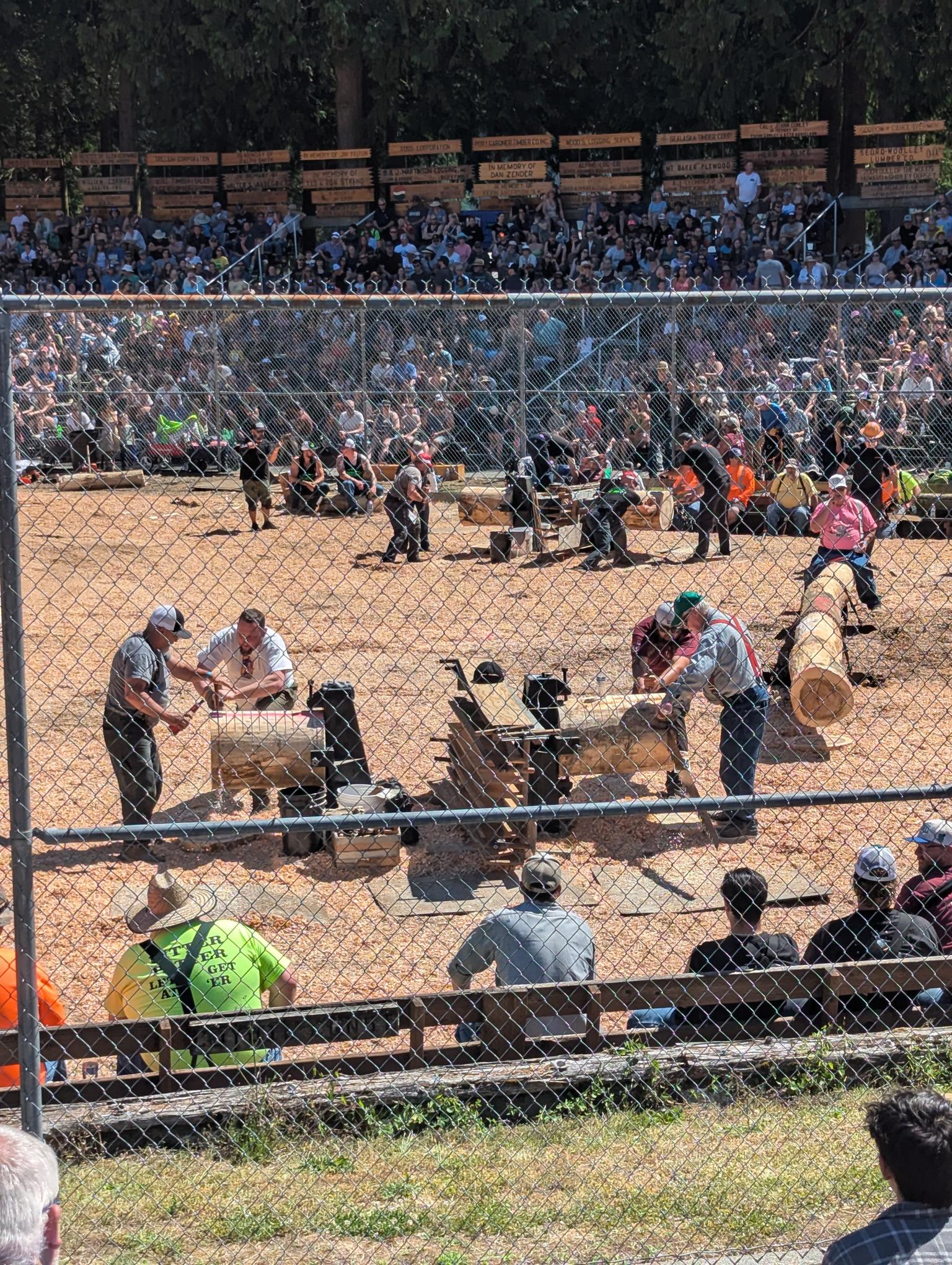 Large crowd in bleachers behind a chain-link fence at an outdoor event, with workers on the dirt arena in the foreground.