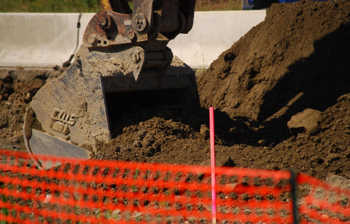 Excavator bucket digging into soil at a construction site with an orange safety barrier in the foreground.