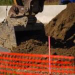 Excavator bucket digging into soil at a construction site with an orange safety barrier in the foreground.