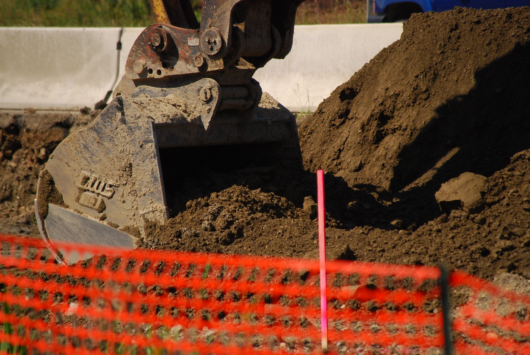 Excavator bucket digging into soil at a construction site with an orange safety barrier in the foreground.